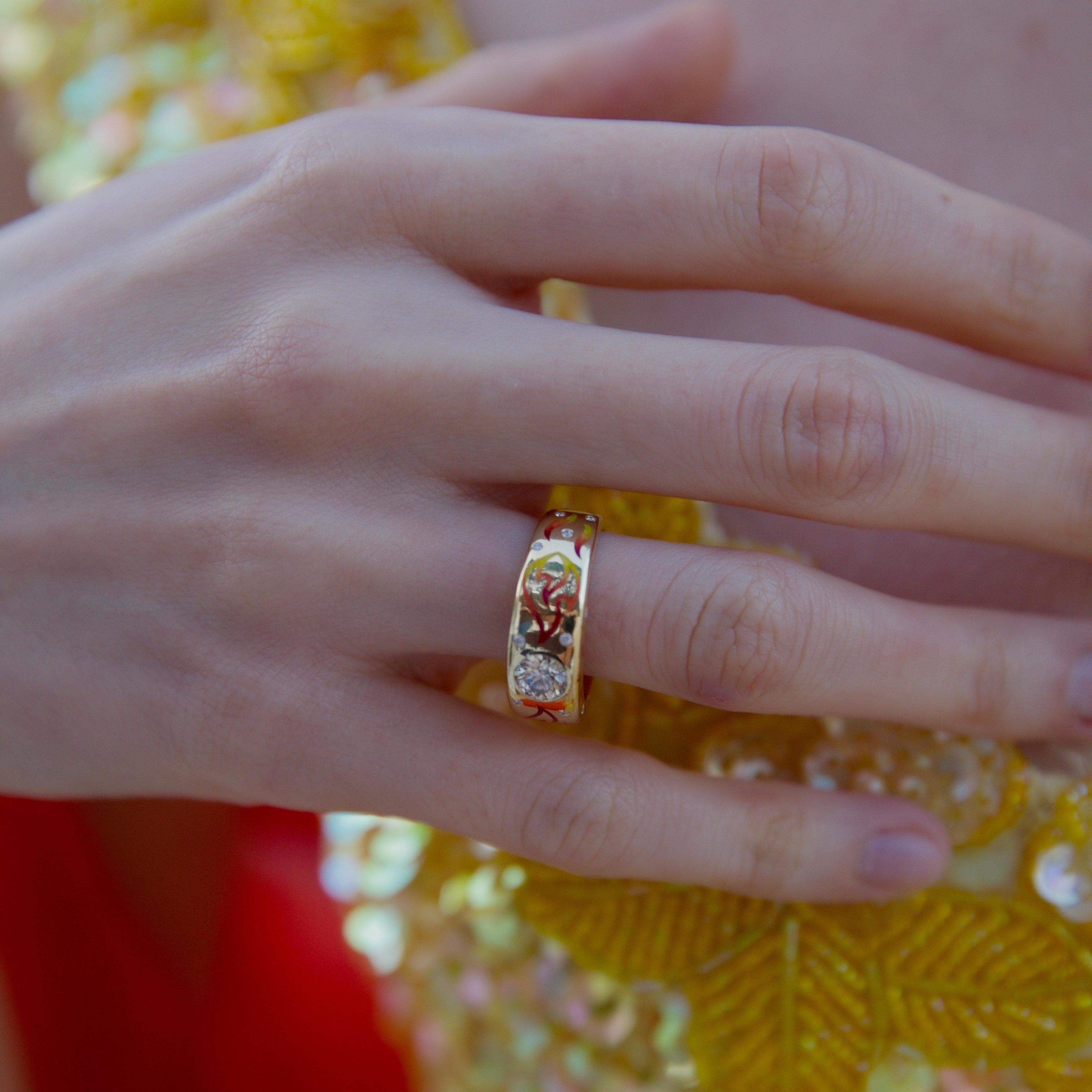Close-up of a hand wearing a colorful ring with a yellow sequined garment in the background.
