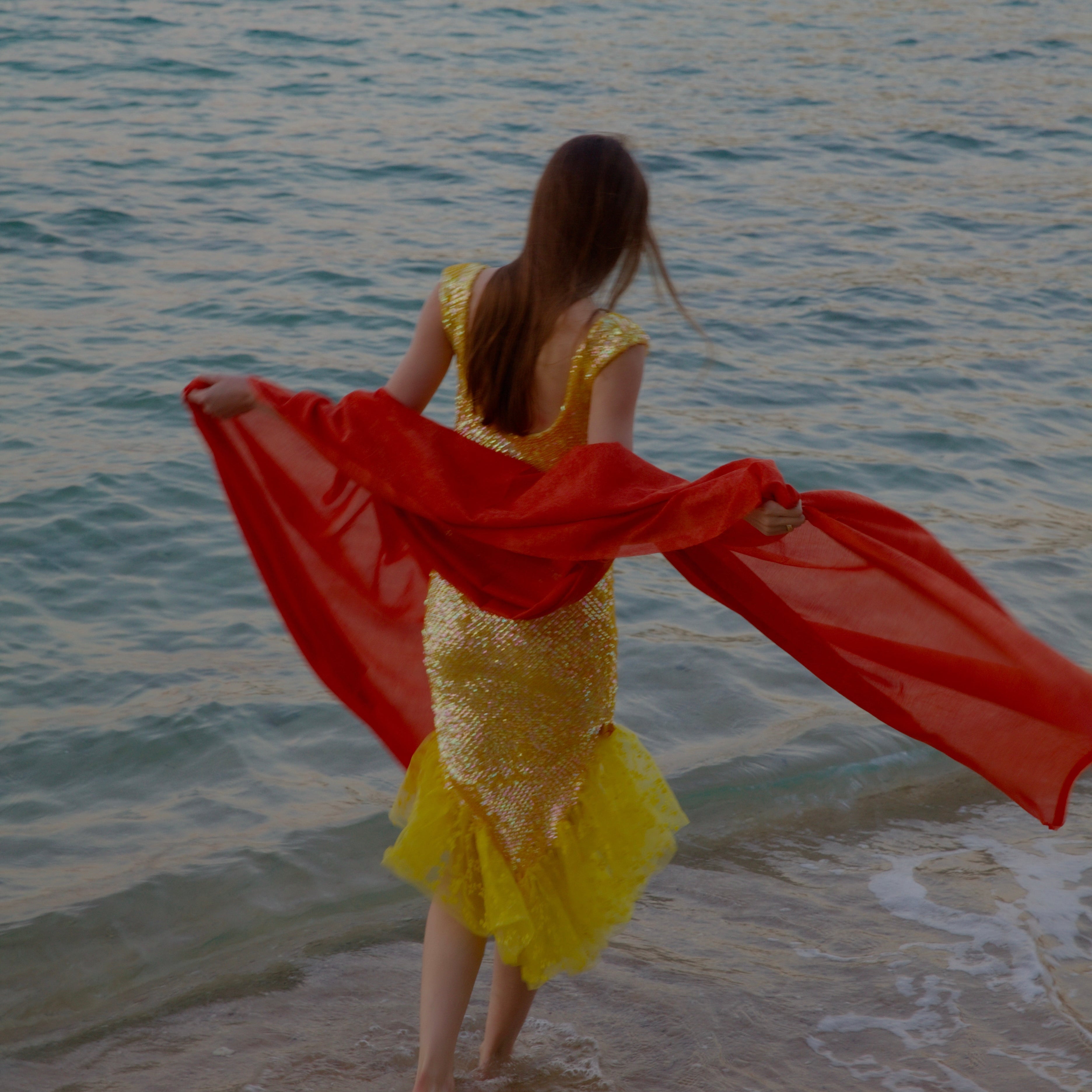 Woman in a yellow dress with a red scarf standing on a beach by the water.