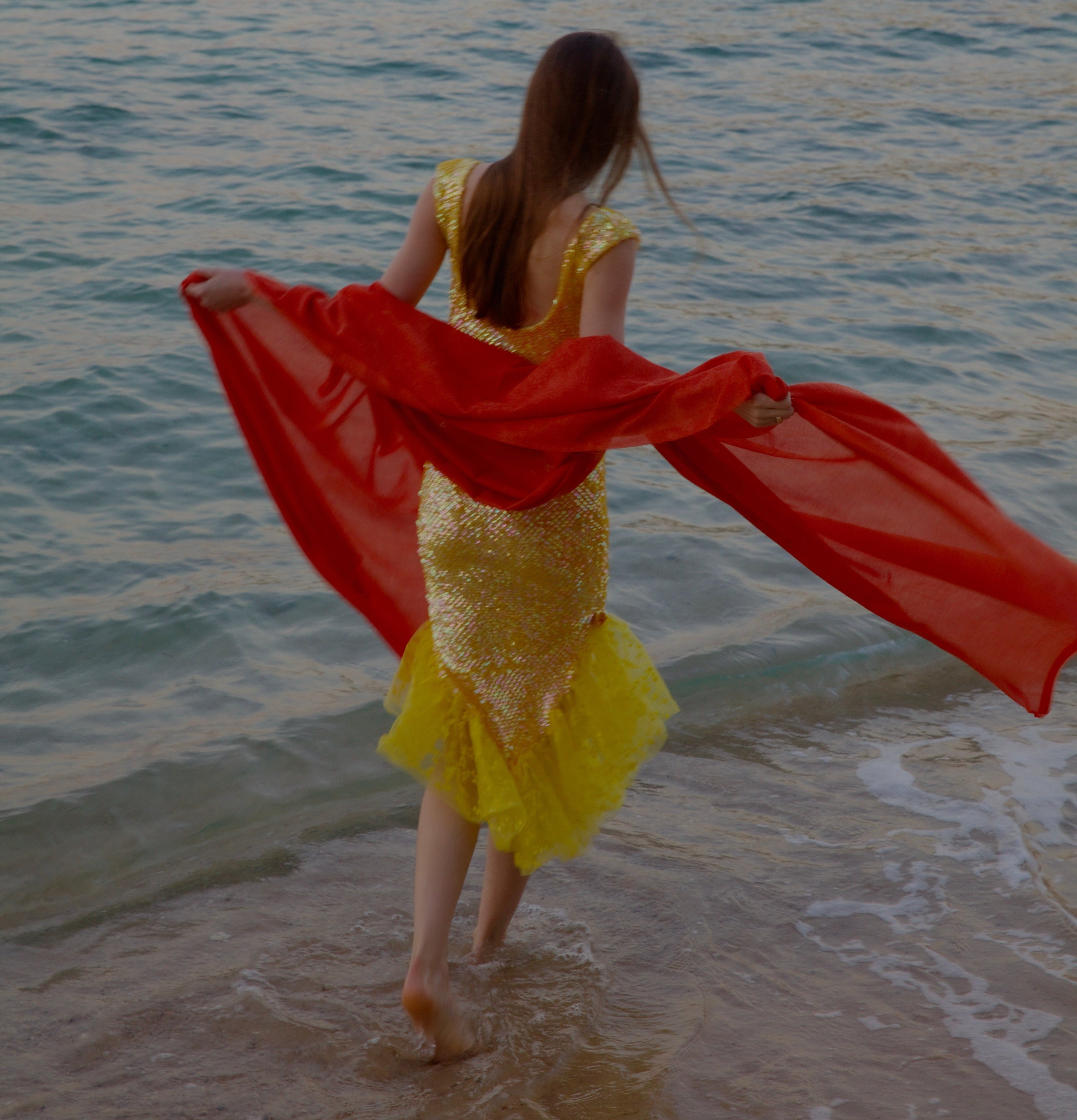 Woman in a yellow dress with a red scarf standing on a beach by the water.
